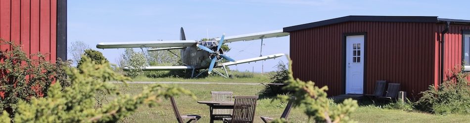 Ett gammalt militärt flygplan står parkerat på Bunge flygfält vid Bunge Vandrarhem på norra Gotland. I förgrunden finns ett bord med stolar och röda byggnader som skapar en rustik och lantlig miljö, omgiven av gröna buskar och en klar blå himmel.